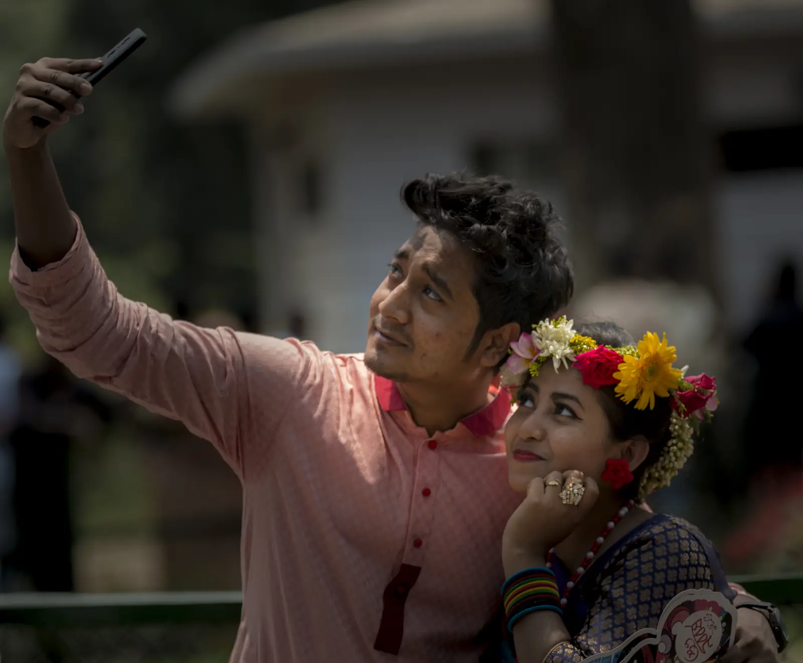 Two people leaning together to take a selfie on a phone held at arm's length, outdoors on a bright day in Bangladesh.